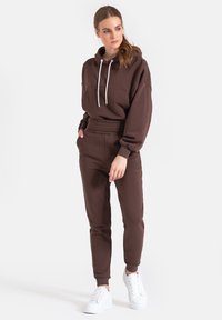 Brązowa bluza z kapturem z białymi sznurkami, połączona z dopasowanymi dresami ze ściągaczem w pasie, noszone z białymi trampkami.