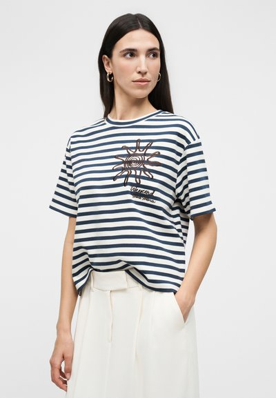 Γυναίκα που φοράει μπλε ναυτικό και λευκό ριγέ μπλουζάκι με γραφικό ήλιου και λευκό παντελόνι ψηλόμεσο, στέκεται με το ένα χέρι στην τσέπη.