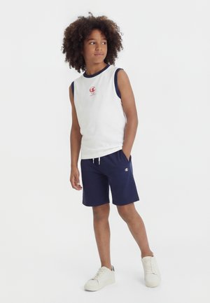 Αγόρι με σγουρά μαλλιά που φοράει λευκή μπλούζα χωρίς μανίκια και σκούρο μπλε σορτς, στέκεται άνετα με το ένα χέρι στην τσέπη.