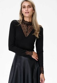 Czarny top z długim rękawem z przezroczystymi koronkowymi akcentami przy dekolcie i mankietach, w połączeniu z czarną skórzaną spódnicą. Gładka faktura na całej powierzchni.