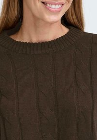 Ciemnobrązowy sweter w warkocze z ribbowanym okrągłym dekoltem. Tekstura jest miękka i gruba, z wystającymi wzorami warkoczy biegnącymi pionowo.