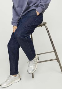 Donkerblauwe denim jeans met een rechte pasvorm, gedragen met een grijze hoodie en zilveren sportschoenen. Zittend op een metalen stoel.