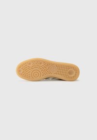 Schuhsohle mit einem beigen Gummibase, die ein strukturiertes Muster, kreisförmige Akzente und ein Wellenmuster für Grip und Traktion aufweist.