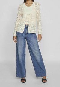 Cárdigan de punto abierto en color crema con mangas largas y cierre de cordón, superpuesto a una blusa ceñida clara, combinado con vaqueros de pierna ancha en color azul.