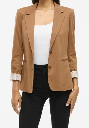 Femme portant un blazer beige avec des poignets rayés retroussés, un haut blanc et un pantalon noir, montrée des épaules aux hanches sur fond blanc.