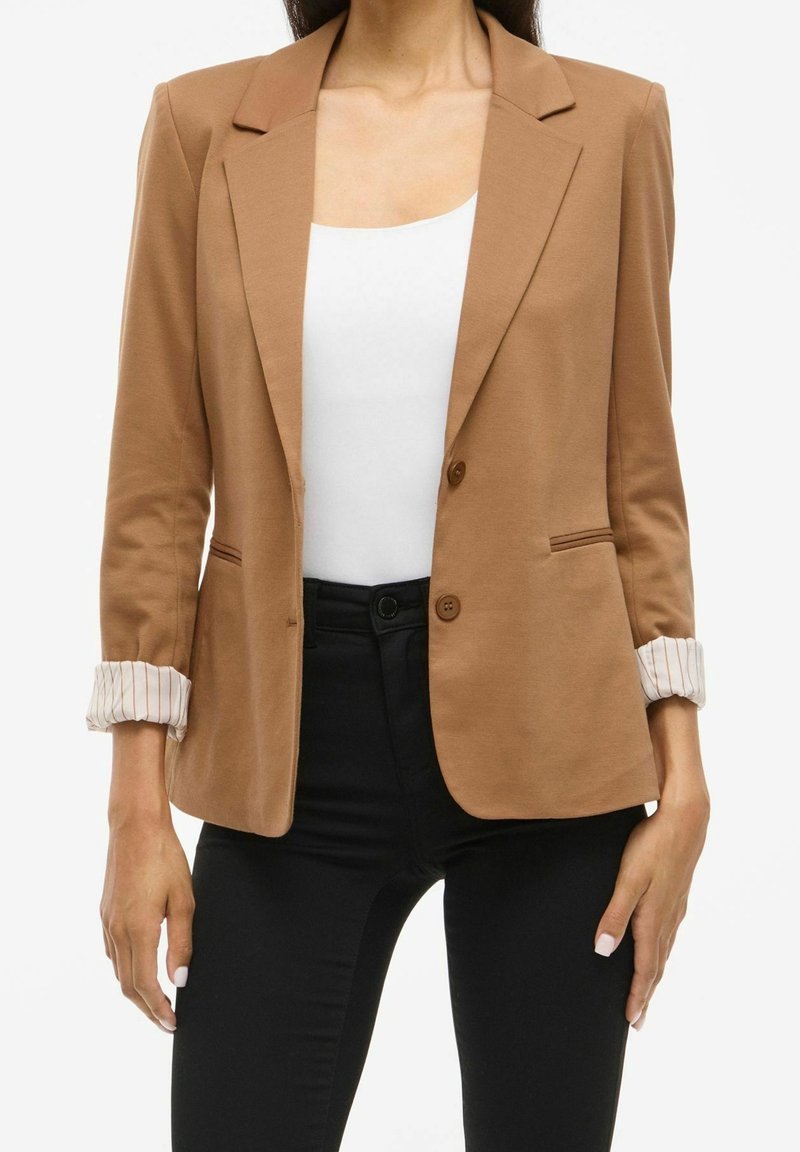 Femme portant un blazer beige avec des poignets rayés retroussés, un haut blanc et un pantalon noir, montrée des épaules aux hanches sur fond blanc.
