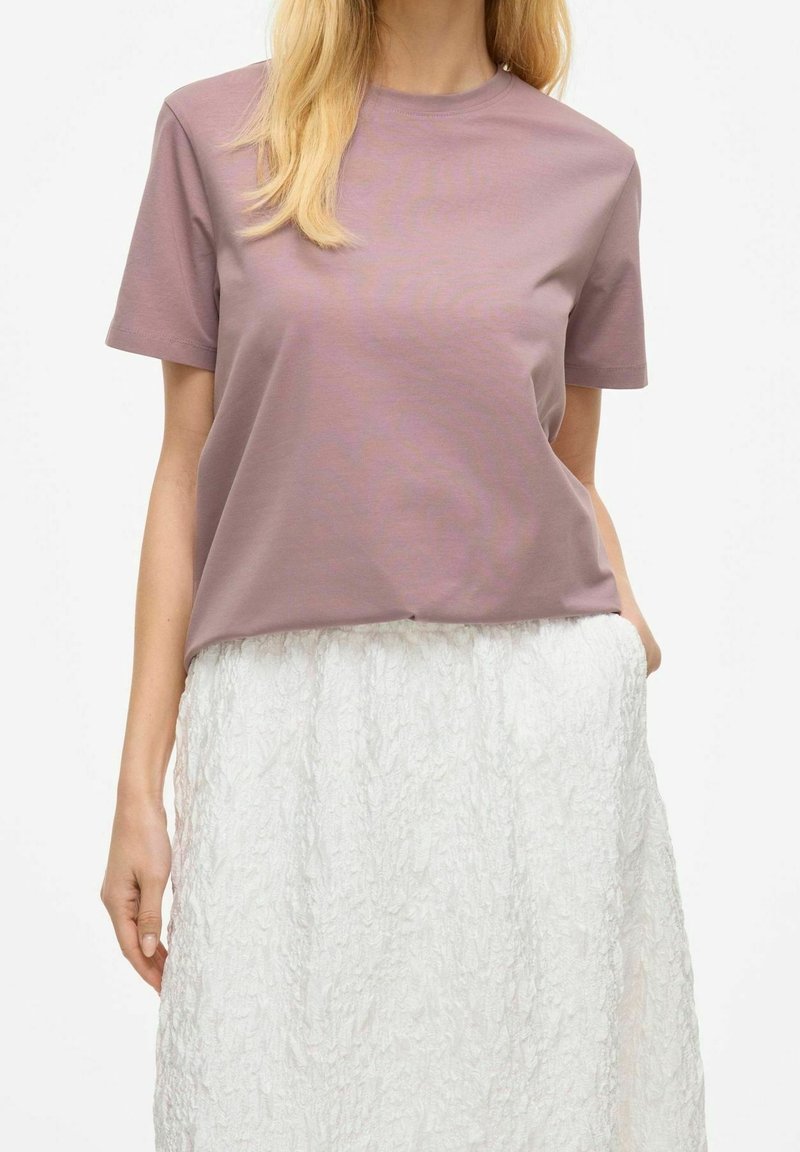 Femme portant un t-shirt mauve à manches courtes rentré dans une jupe blanche texturée aux genoux, debout devant un fond blanc uni.