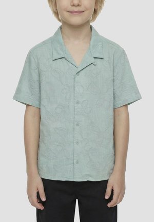 Dijete nosi svijetlozelenu košulju s kratkim rukavima s cvjetnim uzorcima i crne hlače, stoji nasuprot jednostavnoj pozadini.