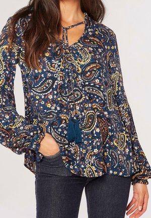 Femme portant un chemisier bleu marine avec un imprimé paisley multicolore et floral, cordons à pompons au col, associé à un jean bleu foncé.