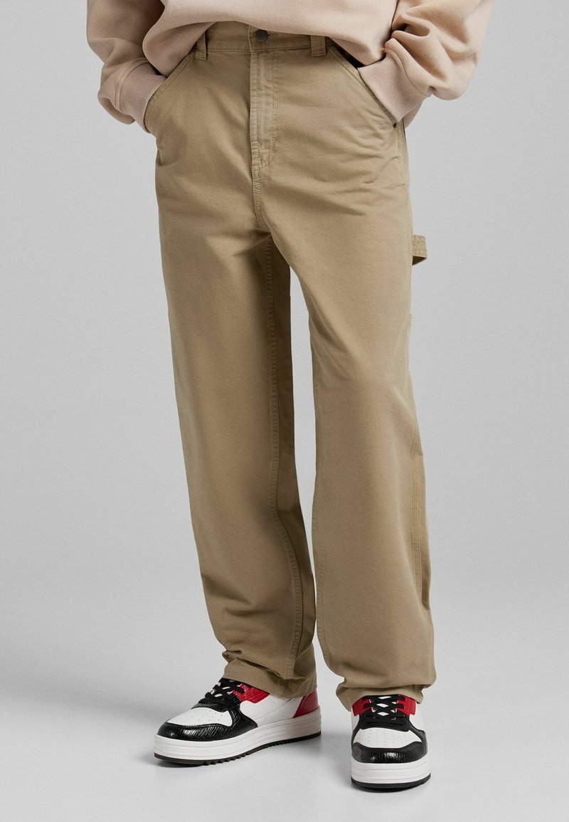 Personne portant un pantalon beige droit avec boucle latérale et des baskets basses blanches, noires et rouges, mains dans les poches, sur un fond gris uni.