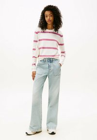 Femme aux cheveux bouclés portant un pull blanc avec des rayures roses, un jean large bleu clair, des baskets blanches et un petit sac à épaule blanc.