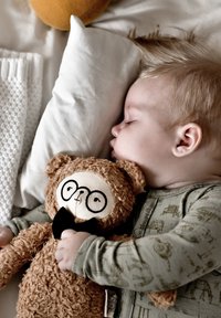 Ursuleț de pluș maro deschis, cu ochelari și papion negru, ținut de un copil adormit îmbrăcat într-un combinezon verde cu model, pe un pat de culoare deschisă.