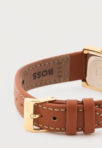 Καφέ δερμάτινο λουράκι ρολογιού με μπεζ ραφές, αγκράφα σε χρυσαφί απόχρωση με ανάγλυφη επιγραφή BOSS και ανάγλυφο λογότυπο BOSS στην εσωτερική πλευρά του λουριού.
