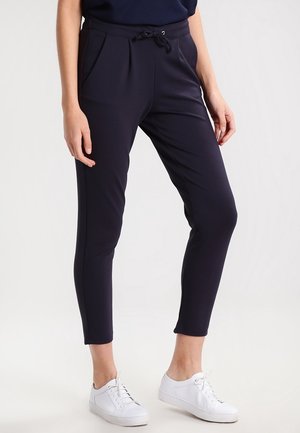 Person trägt dunkelblaue Slim-Fit-Hosen mit Seitentaschen und weiße Sneakers, steht vor einem schlichten hellen Hintergrund.