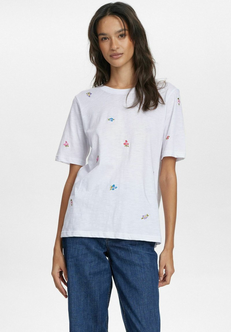 Camiseta blanca de manga corta con bordados florales coloridos dispersos. Textura suave, ajuste relajado, diseño de cuello redondo. Combinada con unos vaqueros azules.