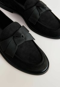 Schwarze Wildleder-Slipper mit einer markanten schwarzen Schleife vorne, einer eleganten, abgerundeten Zehenpartie und strukturierten Sohlen-Details.