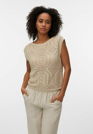 Žena s kučeravými vlasmi oblečená v béžovom bezrukávovom háčkovanom topíku a svetlobéžových nohaviciach s rukami v vreckách, stojaca pred jednoduchým pozadím.