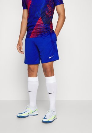 Homme portant un maillot de sport bleu et rouge, un short Nike bleu, des chaussettes hautes blanches Nike et des crampons Nike blancs et bleus, debout avec une main dans la poche.