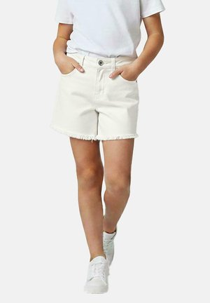 Persoon draagt witte denim shorts met rafelige zomen, wit t-shirt en witte sneakers, handen in zakken, lopend naar voren.