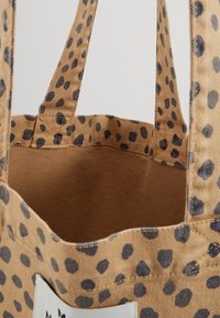 Tote-väska i tan-färgad tyg med svarta oregelbundna fläckar, två handtag och en öppen topp som visar det tomma inre.