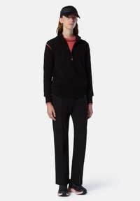 Maglione nero con zip e accenti rossi, abbinato a pantaloni neri e sneakers nere con suole rosa. Il cappello è nero con un design elegante.