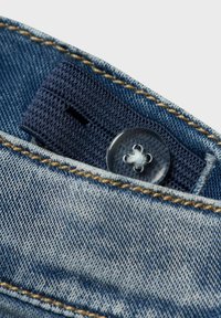Närbild av denimjeans med ett mörkblått vävt bälte, en tydlig knapp med korsstygnsdetalj och gyllene sömmar runt midjan.