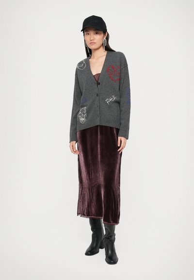 Grijze gebreide cardigan met verschillende borduursels, gedragen over een bordeaux fluwelen slipdress, gecombineerd met zwarte kniehoog laarzen en een zwarte pet.