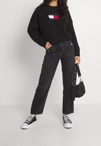 Person som bär en svart Tommy Jeans-tröja, svarta högmidjade byxor, svarta högskor, och håller en svart handväska med en nitarrem.