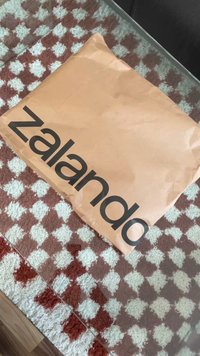 Rudas popierinis voke su juodu užrašu "Zalando". Jis padėtas ant raštuoto kilimo su pakaitomis raudonais ir baltais kvadratėliais.