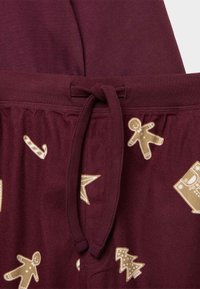 Pantalones burdeos con cintura ajustable, que presentan estampados de temática navideña en beige, incluyendo hombres de jengibre, bastones de caramelo y árboles. Textura suave.