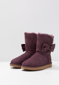 Paire de bottes d'hiver en daim violet avec doublure en peau de mouton et nœud décoratif sur le côté, semelles en caoutchouc marron clair, posées sur une surface blanche.