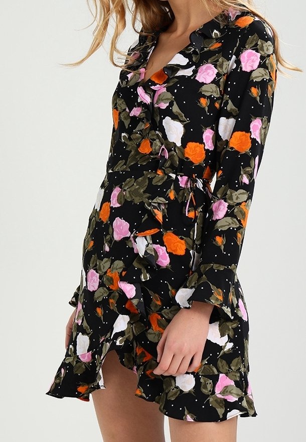 Femme portant une robe portefeuille noire à fleurs avec manches longues et ourlet à volants, avec des fleurs roses, orange, blanches, et des feuilles vertes.