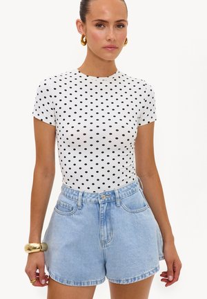 Vrouw draagt een wit kortemouwtopje met zwarte polkadots, lichtblauwe high-waisted denim shorts, gouden creolen en een gouden armband.