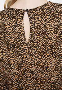 Bakvy av en blus med svart och orange prickmönster, med en tårformad nyckelhålshalsring som är fastsatt med en enda svart knapp.