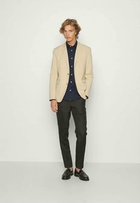 Beiger Blazer mit einem Knopf, marineblaues Hemd, schwarze Hose und schwarze Schuhe. Glatte Texturen, taillierte Passform und schlichte Gestaltung.
