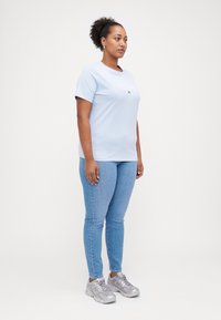 T-shirt bleu clair à manches courtes avec un col rond et un petit logo. Associé à un jean bleu ajusté et des chaussures de sport argentées.