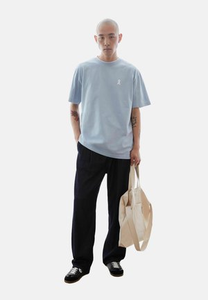 Man with tattoos wearing a light blue t-shirt, black pants, and black sneakers, holding a beige tote bag in his right hand.