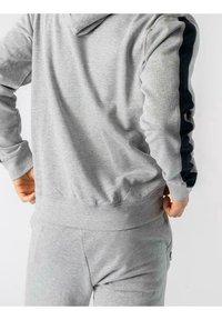 Šedá mikina s kapucí, tmavě modré boční pruhy, žebrované manžety a pas. Vyrobena z měkké bavlněné směsi, s volnějším střihem.