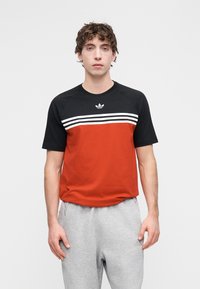 Camisola de manga curta preta e vermelha com riscas brancas ao peito e um logótipo da Adidas. Apresenta decote redondo e mangas raglan.