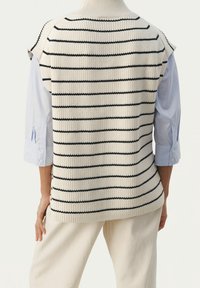 Person, der einen cremefarbenen gestrickten ärmellosen Pullover mit schwarzen horizontalen Streifen über einem hellblauen Langarmshirt und cremefarbenen Hosen trägt, mit dem Rücken zur Kamera steht.