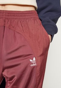 Gros plan sur une personne portant un pantalon de survêtement Adidas bordeaux avec une ceinture élastique et la main dans la poche droite, assorti à une manche de sweat-shirt bleu marine.