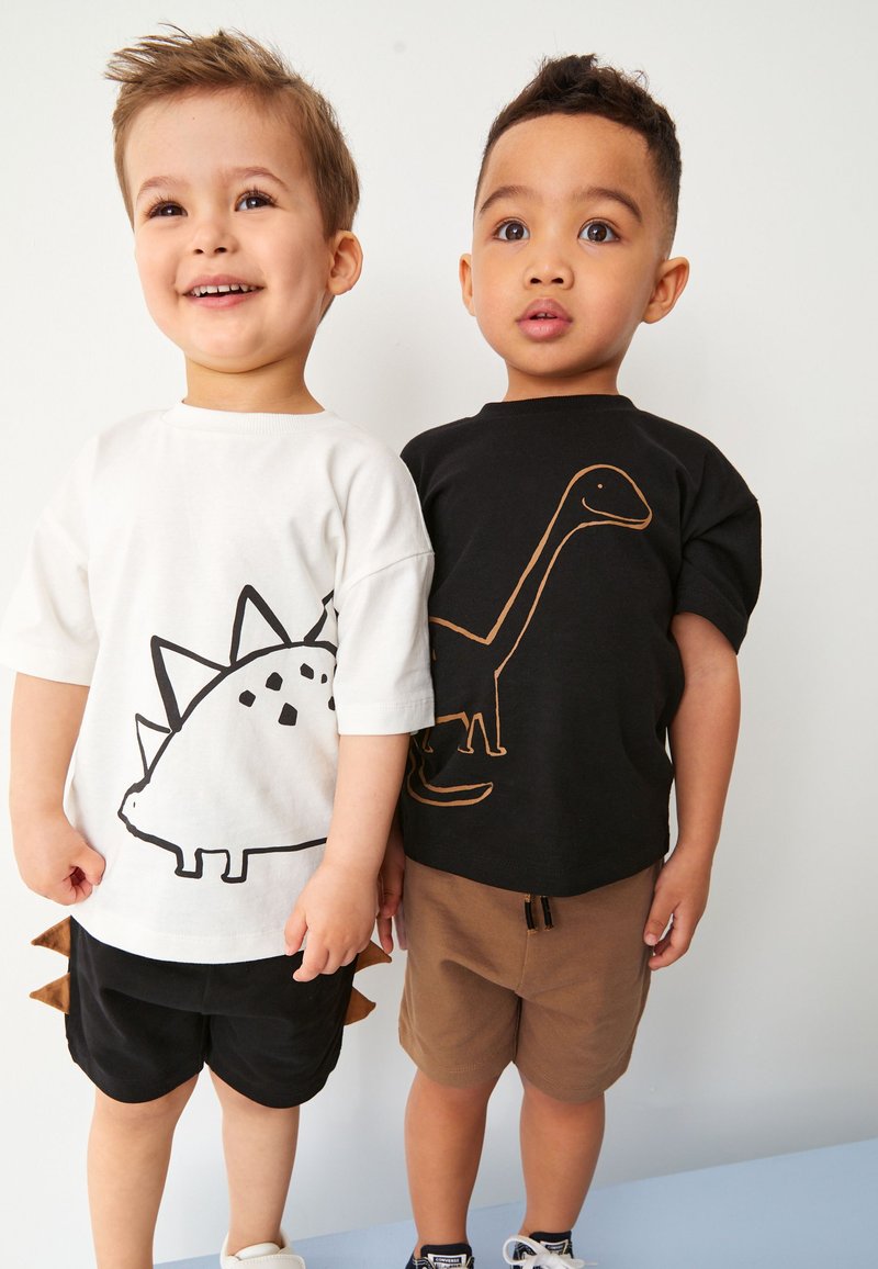 Deux jeunes garçons se tiennent côte à côte ; l’un porte un t-shirt blanc avec un stégosaure et un short noir avec des pointes sur la queue, l’autre un t-shirt noir avec un dinosaure au long cou et un short marron.