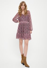 Bloemenjurk met lange doorzichtige mouwen, een getailleerde taille en een gerimpelde zoom. Kleuren zijn donkerroze, paars en groen. Draagbaar met tan enkellaarsjes.