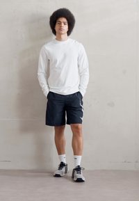 Chemise blanche à manches longues, shorts noirs, baskets grises et chaussettes blanches. Pose debout avec un fond uniforme et un éclairage naturel.