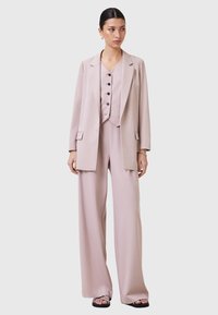 Completo tre pezzi rosa chiaro con giacca lunga, gilet con bottoni e pantaloni a gamba ampia. Tessuto liscio con dettagli minimi.