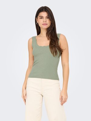 Femme aux longs cheveux bruns portant un débardeur côtelé vert sauge et un pantalon crème, debout devant un fond clair uni.
