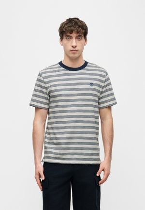 Jeune homme portant un t-shirt à manches courtes beige et bleu marine à rayures et un pantalon noir, debout devant un fond clair uni.
