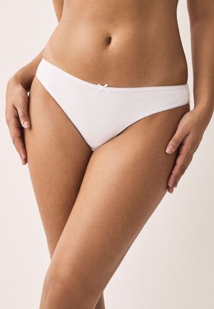 Mujer vistiendo ropa interior blanca sencilla estilo bikini con un pequeño detalle de lazo, con las manos descansando sobre sus caderas, sobre un fondo neutro.