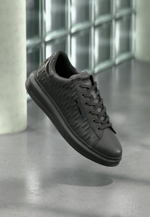 Baskets en cuir noir avec motifs embossés et lacets, de marque "Karl Lagerfeld", présentées flottant au-dessus d'un sol gris près de blocs de verre.