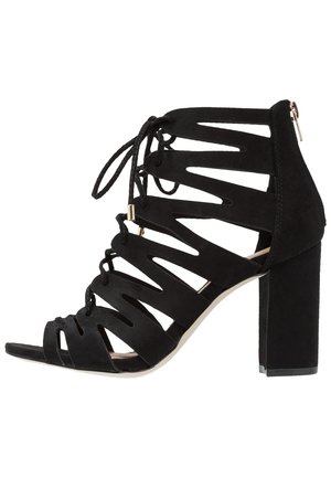 Sort sandal med høj hæl i ruskind med åben tå, zigzag-udskæringer, snørebånd foran og lynlås bagpå på blokhæl.